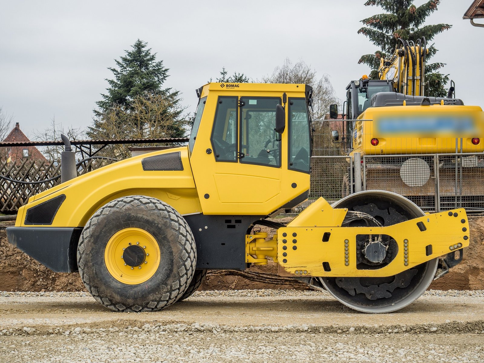 Rouleau compacteur monocylindre Bomag vu de profil sur une couche de gravier, une pelle en arrière-plan